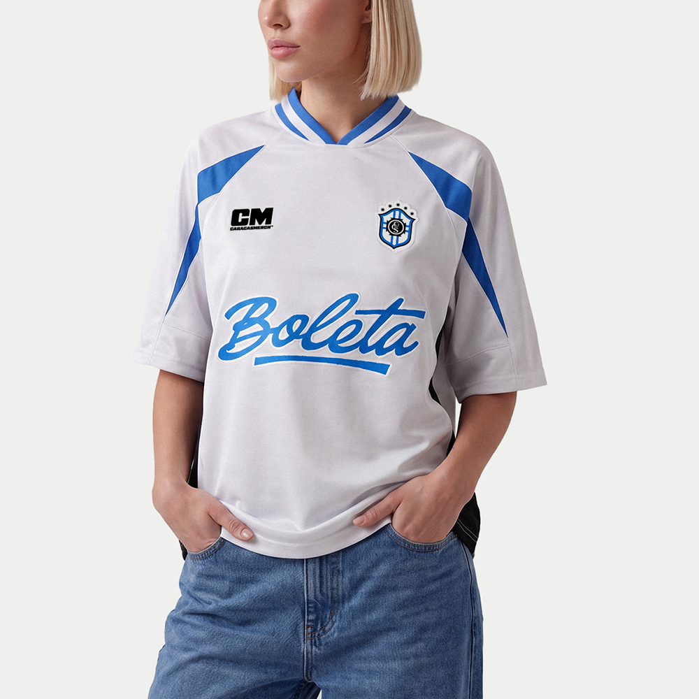 Mujer llevando la camiseta de fútbol boxy blanca y azul «Boleta» de Caracas Merch con vaqueros